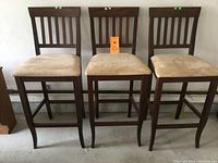 Three wooden counter height stools with padded beige suede look seats, slatted backrest design, and square bases measuring 17 inches. The seats have a height of 29.5 inches. Condition shows usage marks on wood.