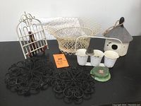 Overview of all items in the lot arranged on table: wire basket, garden gate display, birdhouse, two metal trivets, two flower frogs, and a card