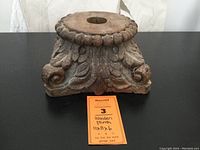 Front view of square wooden plinth with carved leaf and scroll detailing and beaded top edge showing hole in center and loose curl secured by nail.