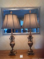 Two brass tone antique style table lamps with silk shades on a wooden surface in front of a silver-bordered mirror.
