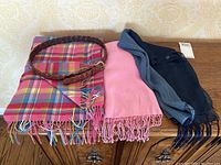 Photo shows a pink pashmina scarf, a multicolor plaid scarf, a two-toned blue scarf and a braided brown leather belt laid out on a wooden surface.
