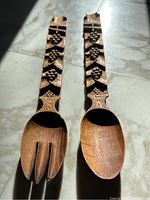 Side by side carved wooden serving spoon and fork showing detailed grape and leaf carving along the handles and smooth wooden spoon bowl and fork tines.