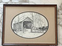 Framed vintage black and white oval-shaped print of covered bridge with horses and carriage. Print is signed and numbered and in good condition.