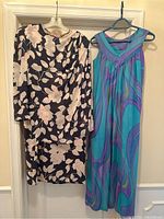 Two vintage dresses hanging against a door; one black with pink rose print and buttons on shoulder, the other sleeveless with a blue and purple abstract print.