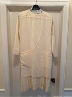 Front view of vintage cream wool Louis Féraud dress hanging on hanger, showing long sleeves, round neckline, front pockets, and pleated bottom hem.