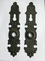 Photo of two antique ornate metal door back plates with decorative detailing, showing full length on white background.