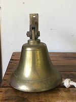 Front view of large brass bell showing smooth body with patina and mounting attachment on top, including white rope pull