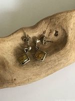 Pair of small sterling silver earrings with square Baltic amber stones placed on a carved wooden surface.