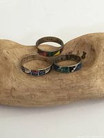 Three sterling silver enamel rings with colorful rectangular enamel patterns on silver bands. Rings displayed on natural wood piece. View shows tarnishing and surface wear.