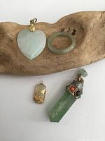 Photo showing four jewelry items on a textured wooden surface including two jade pieces, a small gold-tone pendant, and a crystal pendant with colored stones.