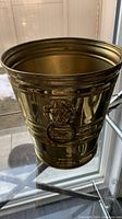 Front view of antique solid brass trash can showing lion head doorknocker handles on each side and polished surface.