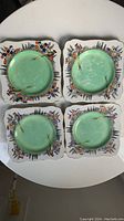 Four vintage square dessert plates arranged flat, showing green central glaze and floral bird designs along square scalloped edges.