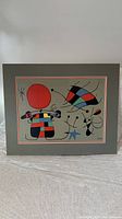 Frontal view of the signed Joan Miró silkscreen painting with abstract shapes and a large red circle on a gray background with a signature.