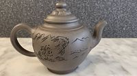 Side view of brown Yixing Zisha clay teapot showing engraved mountain and house landscape on body and curved spout and handle.