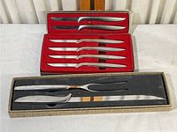 Full view of steak knives set in a black box with red velvet lining and carving knife with fork in a separate beige textured box.