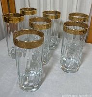 Seven clear tall glasses with ornate gold-colored rim, showing pattern detail and height relative to table surface.