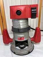 Front view of router showing red plastic top, metal body, dual side handles, and base.