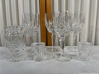 Wide shot of eight Waterford crystal glasses of varying sizes arranged on a white cloth in front of a wooden frame and white curtain.
