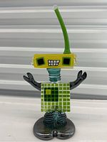 Front view of the colorful art glass robot sculpture showing the yellow head with black eyes, grid-pattern torso, black arms, and metallic base.