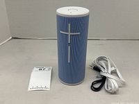 Blue Ultimate Ears BOOM 3 speaker standing upright with volume buttons visible, accompanied by white charging cable and black aux cable coiled beside it on gray surface.
