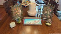 Overview of antique and vintage decorative items including two ornate metal photo frames with floral prints, Carlton Ware vase, glass paperweight, porcelain trinket box, enameled copper dish, and mermaid jewelry holder arranged on wooden table.