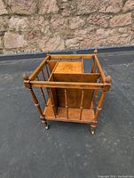 Angled view of burled walnut Canterbury style magazine stand showing square frame, vertical slats, corner finials, and brass casters on a black surface outside.