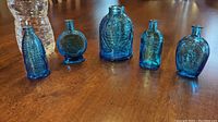Five blue glass Wheaton mini bottles displayed upright on a wooden table with embossed designs and varying shapes.