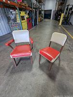 Two vintage Steelcase Tanker chairs pictured side by side on a concrete floor inside a warehouse setting. One chair has arms with red vinyl armrests and seat, cream vinyl backrest, and wooden legs. The other chair is armless with matching upholstery colors and wooden legs.
