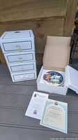 Five white boxes stacked containing individual Christmas lighted plates from Rhodes Studios with one open box showing a hand painted plate inside and paperwork including certificates of authenticity.