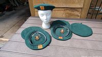 Six vintage green wool Basque beret hats displayed on a table, some placed on a mannequin head, showing varied styles with emblem patches and plain ones.