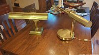 Two heavy brass vintage desk lamps positioned on a wooden table, showing their front and side profiles