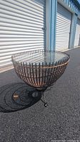 Full view of the wicker fish trap table with glass top on asphalt outside storage units.