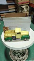 Vintage style yellow and green pickup truck model on a white table in front of a cardboard box