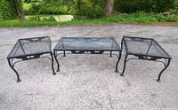 Three vintage wrought iron mesh patio tables, black with paint wear and rust spots visible, arranged side by side outdoors on paved surface