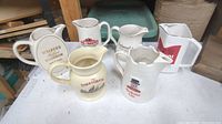 Photo showing six ceramic advertising bar pitchers arranged on a table with wooden shelving in the background. Variations in shape, size, and branding logos visible.