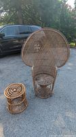 Wicker peacock chair alongside rattan ottoman/table base placed outdoors on pavement with car and trees in background.