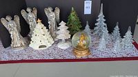Full view of lot showing 3 silver color angel candle holders, white bisque Christmas tree votive holder, green ceramic Christmas tree, 7 clear glass Christmas trees, and gold angel snow globe.