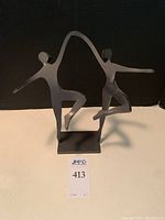 Front view showing two dancing figures connected by a curved arch on a rectangular base with artist signature visible.