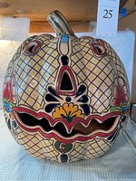 Front view of hollow ceramic pumpkin with carved facial openings and handpainted colorful geometric and floral designs on yellow glazed background with removable brown stem lid.