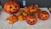 Overall shot showing three orange glass jack-o-lantern decorations in varying sizes alongside orange and red autumn-themed swag with leaves, small pumpkins, and berries on a beige patterned surface.