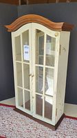 Full view of tabletop cabinet showing wood top, painted cream body with glass doors, and decorative insect accents.