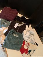 Assorted women’s sweaters in various colors and styles folded on a table, including maroon cable knit, star patterned knits, novelty Santa knit, and button placket sweater.