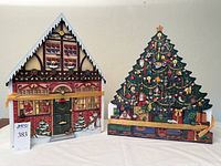 Front view of the two wooden advent houses, one shaped like an alpine chalet and the other a Christmas tree, showing decorative windows, snow, and festive designs