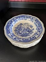 Stack of six blue and white low bowls with scenic rural landscape pattern and floral border on a black surface