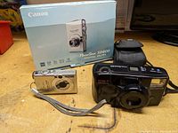 Photo showing Canon PowerShot SD600 camera in silver with original box and Olympus Zoom AF black camera with wrist strap on wooden surface.