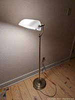 Full view of brass floor lamp powered on showing white curved glass shade casting light on wall.