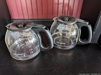 Two Mr. Coffee glass coffee carafes with black plastic handles and lids placed side by side on black surface with red textured background.