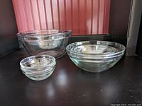 Photo shows a set of seven clear glass mixing and prep bowls of varying sizes, arranged on a dark surface against a red vertical panel background.