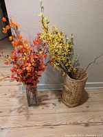 Lot display showing two artificial flower arrangements; orange flowers with dark berries in clear glass vase and yellow flowers in a woven basket.