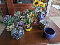 Photo showing all flowers in various pots and vases plus a glass fishing float on a wooden table.